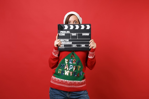Zszokowany młoda dziewczyna Santa ukrywa, zakrywa twarz klasycznym czarnym filmem, co clapperboard na białym tle na jasnym czerwonym tle. Szczęśliwego nowego roku 2019 celebracja party wakacje koncepcja. Makieta miejsca na kopię.