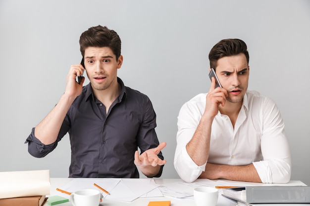 Zszokowany zdezorientowany dwóch młodych biznesmenów współpracujących rozmawiających przez telefony komórkowe.