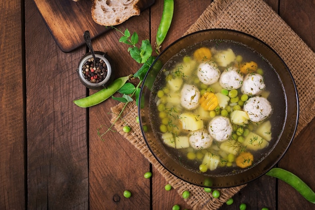 Zdjęcie zupa dietetyczna z klopsikami z kurczaka i groszkiem zielonym w szklanej miseczce na drewnianym tle.