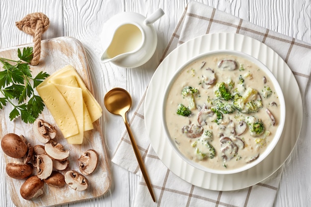Zupa serowa z grzybów brokułowych w białej misce ze złotą łyżką i składnikami na białym drewnianym stole, poziomy widok z góry, zbliżenie