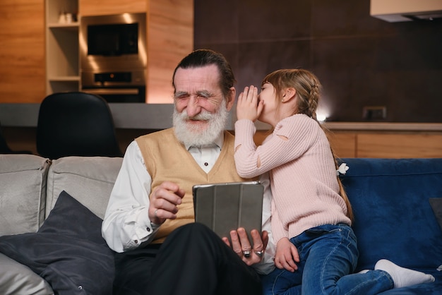 Zwolnione tempo słodkiej radosnej dziewczyny ze śmiesznymi warkoczami, które szepczą jej do ucha dziadka jej sekret, gdy siedzą razem na kanapie w domu