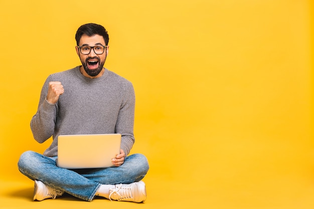 Zwycięzca sukcesu! Portret szczęśliwy młody brodaty mężczyzna za pomocą laptopa i świętuje sukces lub zwycięstwo na białym tle na żółtym tle.