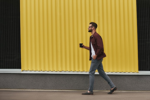 Życie w mieście. Widok z boku przystojnego młodego brodatego mężczyzny w casual i okulary trzymając jednorazowy kubek podczas spaceru ulicą miasta. Styl miejski