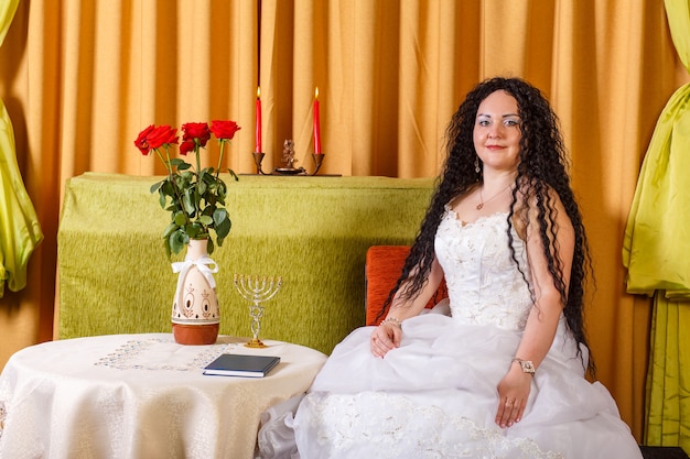 Żydowska panna młoda w białej sukni ślubnej bez welonu siedzi przy stole z kwiatami przed ceremonią chuppy