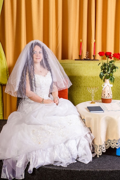 Żydowska panna młoda w białej sukni, z twarzą zakrytą welonem, siedzi przy stole z kwiatami przed chupą. Zdjęcie pionowe