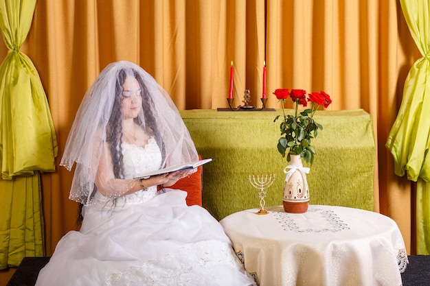 Żydowska panna młoda w białej sukni z zasłoniętą twarzą siedzi i czyta Tegilim przed ceremonią chupa