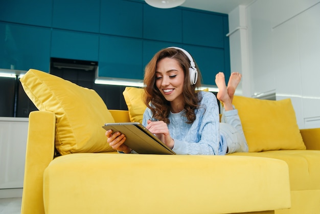 Żywiołowa szczęśliwa kobieta korzysta z komputera typu tablet, leżąc na wygodnej żółtej kanapie i słucha muzyki za pomocą słuchawek.