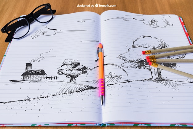 Cuaderno con dibujo de paisaje, lápices y gafas