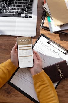 Maquette de demande de visa dans un smartphone tenu par des mains humaines sur un bureau avec passeport, ordinateur portable et documents