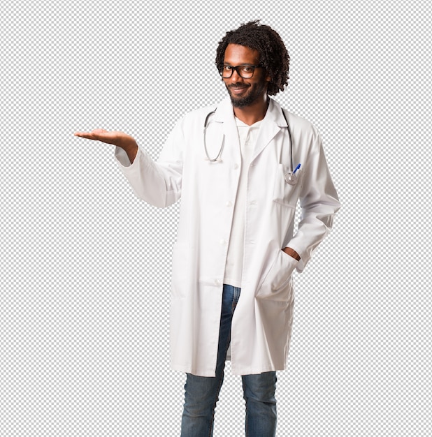 Apuesto médico afroamericano sosteniendo algo con las manos, mostrando un producto, sonriente y alegre, ofreciendo un objeto imaginario
