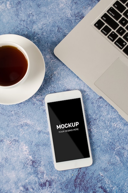 Blanco smartphone con pantalla en blanco negro en el escritorio de oficina con ordenador portátil y una taza de té. Mock up de teléfono.