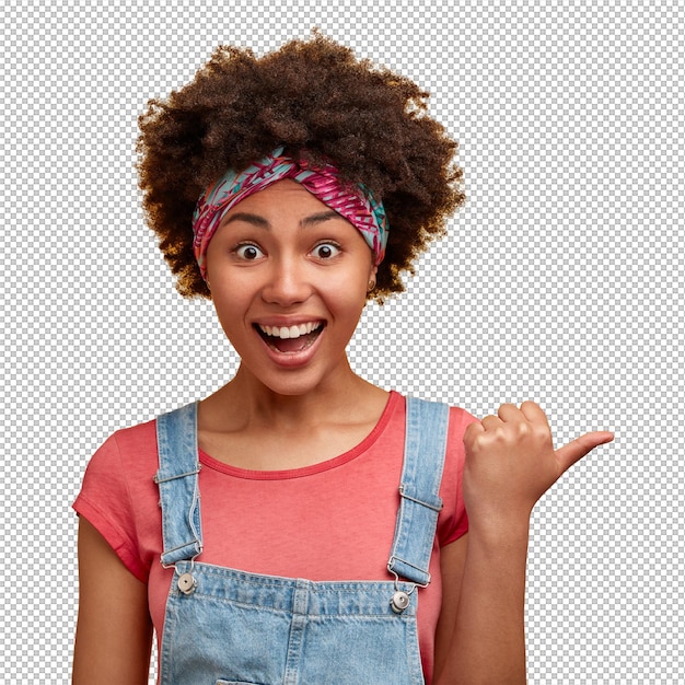 Capture horizontale d'une femme afro-américaine à la peau sombre et bouclée joyeuse avec une expression de joie débordante pointe de côté contre le mur jaune vêtue de dungarees à la mode