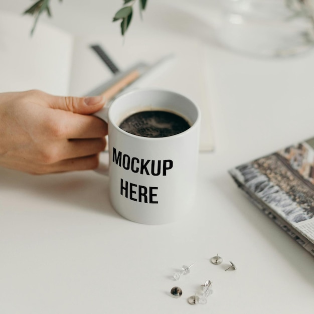 Close-up de uma caneca de café branca com o texto Mockup Aqui mantido por uma pessoa com outros objetos