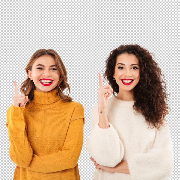 Dos chicas sonrientes en suéteres apuntando hacia arriba sobre la pared blanca
