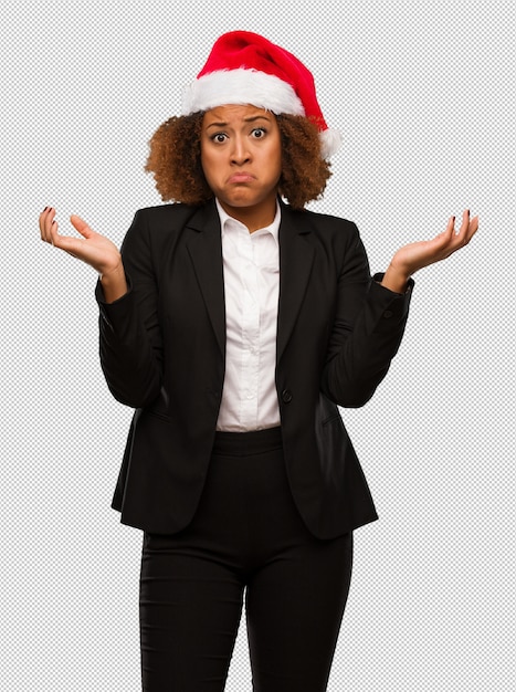 Empresaria negra joven que lleva un sombrero de santa de la Navidad que duda y que encoge hombros