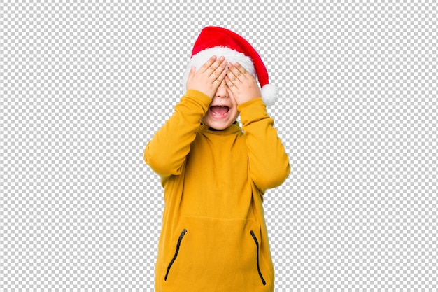 Garotinho, comemorando o dia de natal com um chapéu de papai noel cobre os olhos com as mãos, sorri amplamente esperando por uma surpresa.