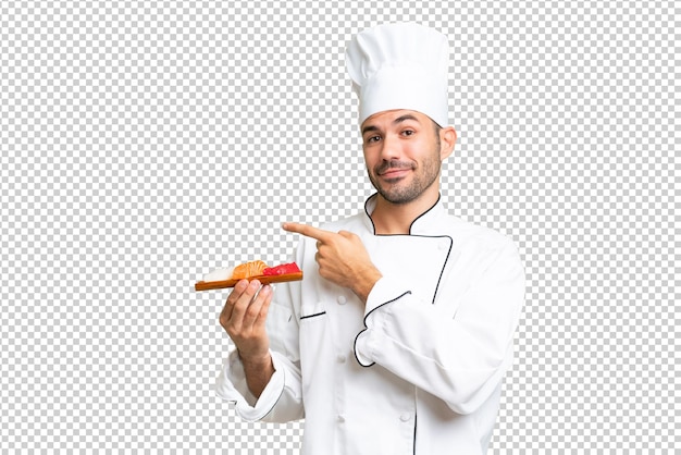 Giovane chef caucasico che tiene un sushi su uno sfondo isolato puntando il dito verso il lato