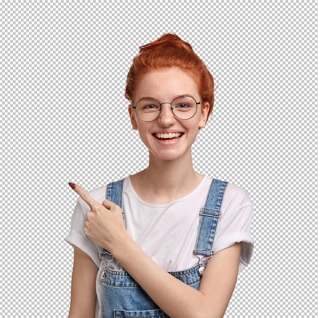 Glückliche frecklige Dame in Brille fühlt sich zufrieden trägt Dungarees zeigt in der oberen linken Ecke an, schlägt vor, den Kopierraum weise zu nutzen, steht gegen die weiße Wand, hat ein breites Lächeln