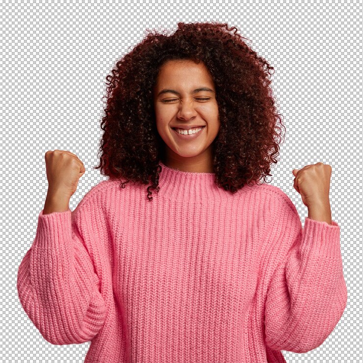 PSD happy dark skinned teenager raises clenched fists celebrates victory and success gained desirable wish smiles broadly dressed in knitted jumper isolated on pink wall