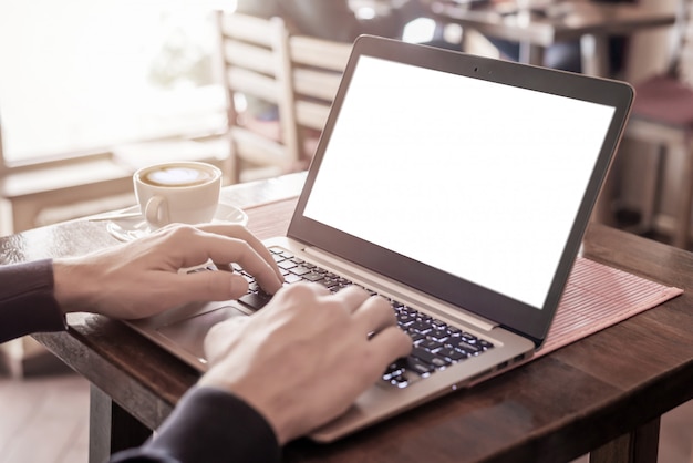 Hombre escribiendo en el teclado del ordenador portátil con pantalla aislada para maqueta. Computadora en la mesa de la cafetería con una taza de café al lado. La luz entra por una ventana