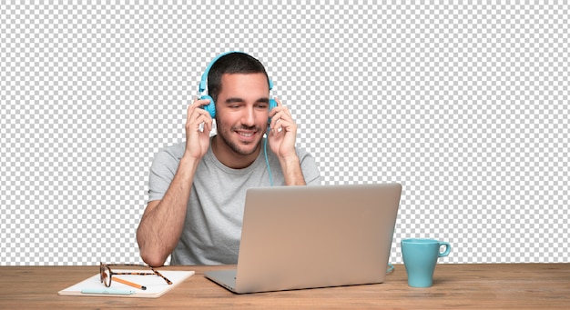 Hombre joven feliz que se sienta en su escritorio y que usa auriculares