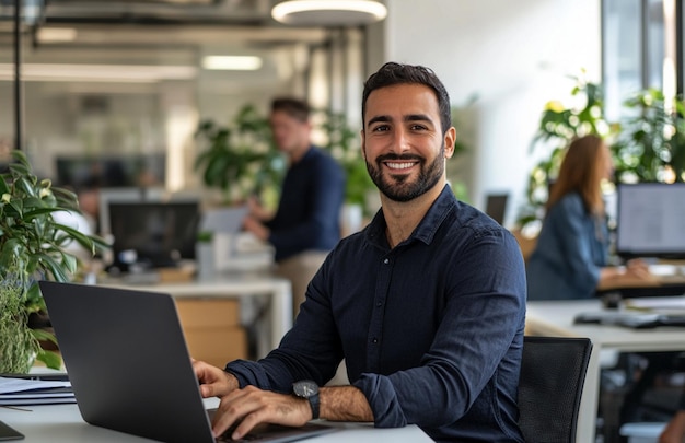 Un hombre de negocios feliz sentado en su escritorio con una computadora portátil y mirando a la cámara mientras está de pie en una oficina