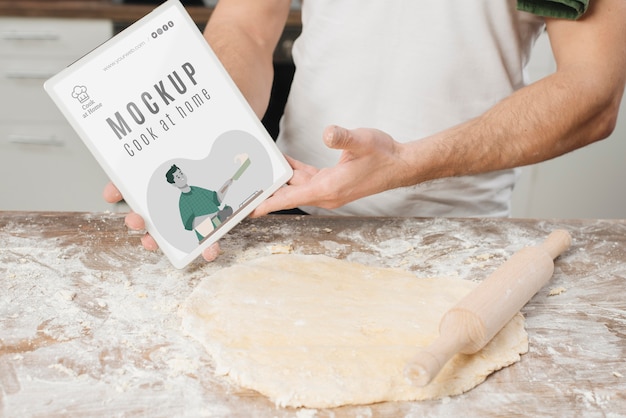 Homem enrolando massa na cozinha e segurando um livro