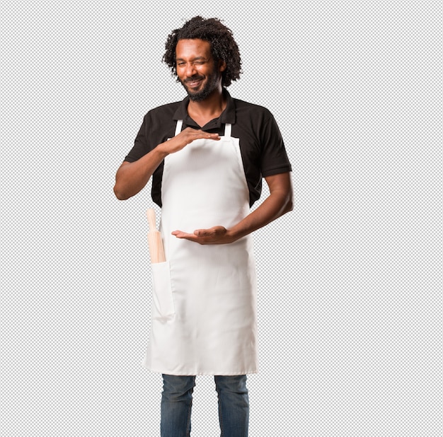 Hübscher Afroamerikanerbäcker, der etwas mit den Händen hält, ein Produkt zeigt, lächelt und nett und bietet einen eingebildeten Gegenstand an