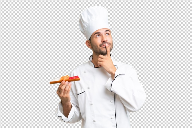 PSD jovem chef caucasiano segurando um sushi sobre um fundo isolado tendo dúvidas enquanto olhava para cima