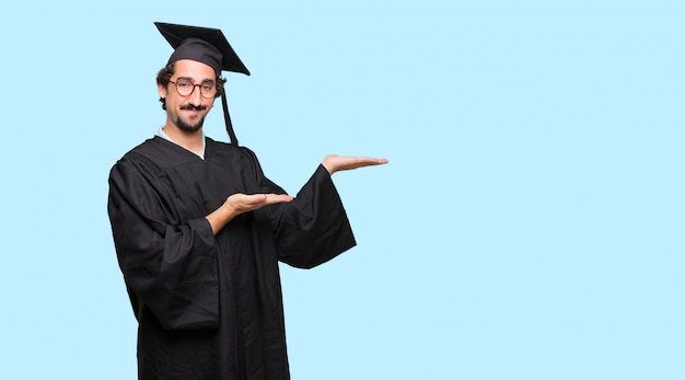 joven graduado sonriendo con una expresión satisfecha mostrando un objeto o concepto con ambas manos