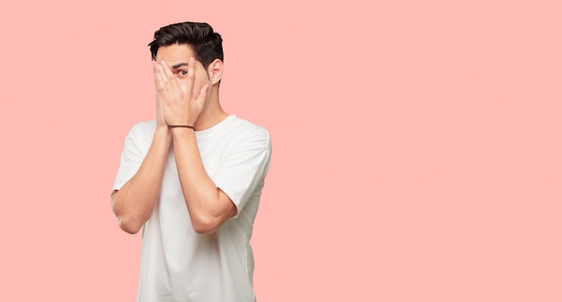 Joven guapo con una expresión asustada y asustada, cubriendo los ojos con ambas manos, mirando