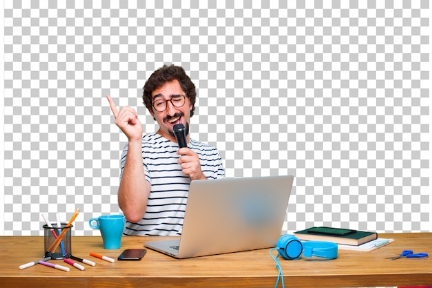 Junger verrückter Grafikdesigner auf einem Schreibtisch mit einem Laptop und mit einem Mikrofon