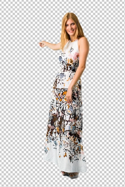 Loira jovem garota em um vestido de verão, apontando para trás com o dedo indicador apresentando um produto