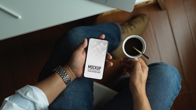 Mann, der das leere Smartphone des Modells verwendet und Kaffeetasse hält, während er im Büroraum sitzt