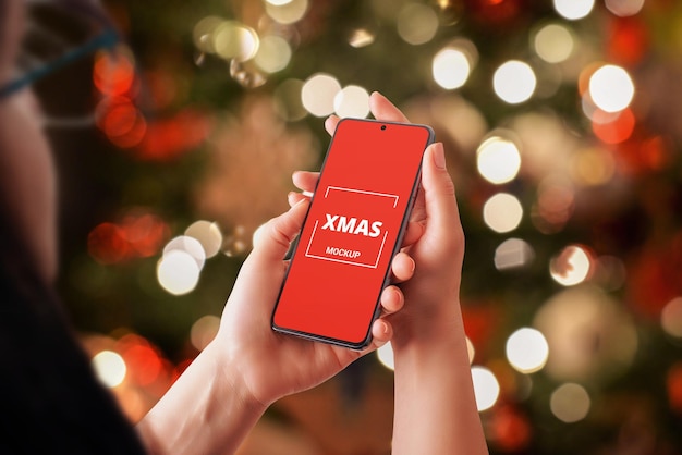 Maquete de telefone nas mãos da mulher com muitas luzes de natal no fundo. tela isolada com smartphone com câmera embutida