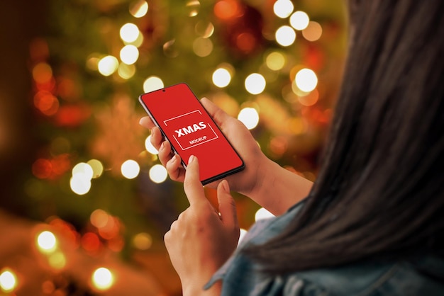 Modello del telefono di Natale in mano della donna. Invio di auguri di Natale per telefono concetto. Luci bokeh di albero di Natale decorato in background