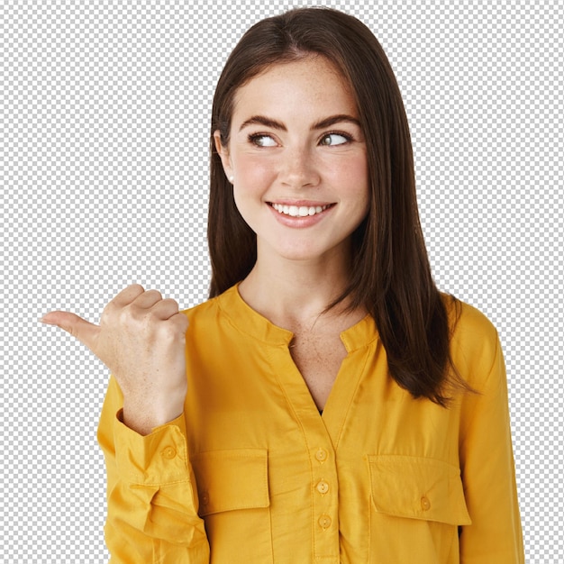 Mujer joven y atractiva comprobando la oferta promocional señalando con el pulgar a la izquierda y mirando fijamente la pancarta con una sonrisa satisfecha de pie en la pared blanca