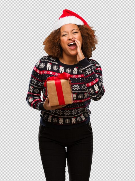 Mujer negra joven que sostiene un regalo en el día de Navidad que grita algo feliz al frente