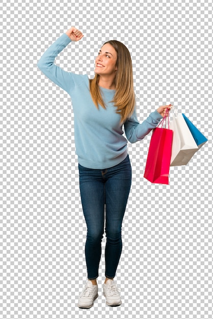 Mujer rubia con la camisa azul que sostiene muchos bolsos de compras en la posición de la victoria