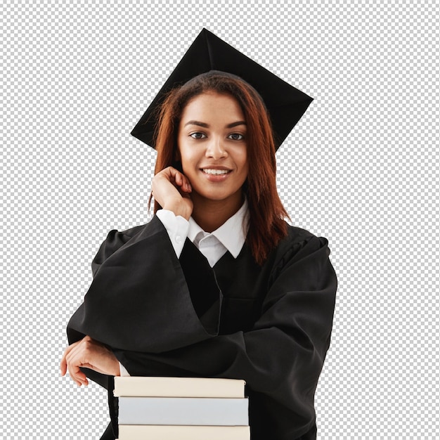 Mulher graduada em boné e manto sorrindo sentada com livros sobre superfície branca