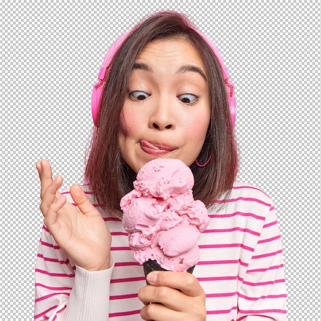 Mulher olha para sorvete apetitoso, lambe lábios, come uma deliciosa sobremesa congelada, ouve música através de fones de ouvido, vestida com um jumper listrado.