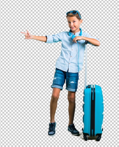 Niño con gafas de sol y auriculares que viajan con su maleta apuntando el dedo hacia un lado.