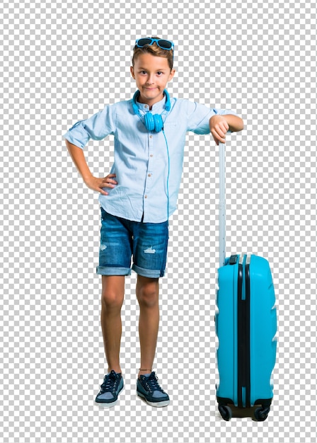 Niño con gafas de sol y auriculares que viajan con su maleta posando con los brazos en la cadera