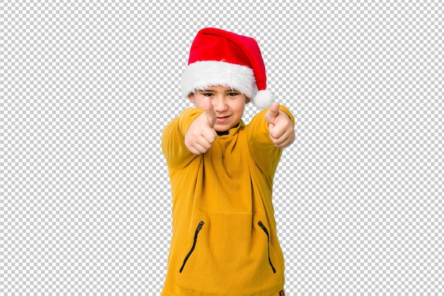 El niño pequeño que celebra el día de Navidad que lleva un sombrero de santa con los pulgares sube, anima sobre algo, apoya y respeta el concepto.