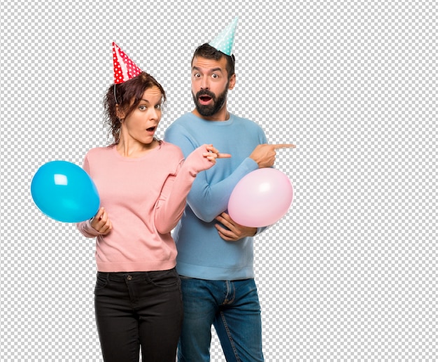 PSD pareja con globos y sombreros de cumpleaños apuntando el dedo al lado con una cara sorprendida