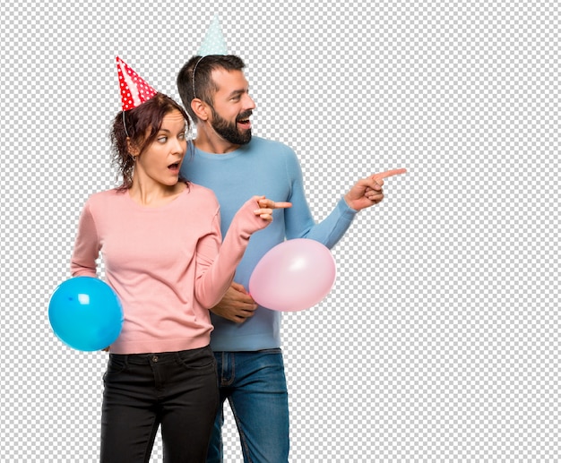 Pareja con globos y sombreros de cumpleaños apuntando con el dedo hacia un lado.