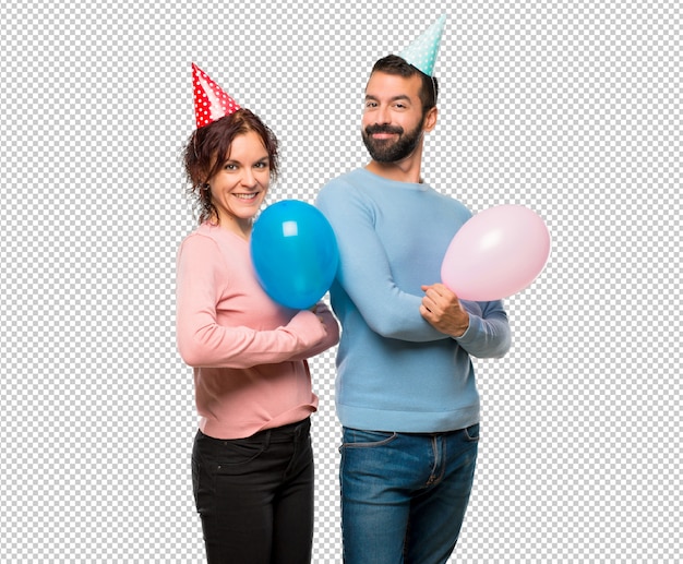 Pareja con globos y sombreros de cumpleaños manteniendo los brazos cruzados en posición lateral mientras sonríe