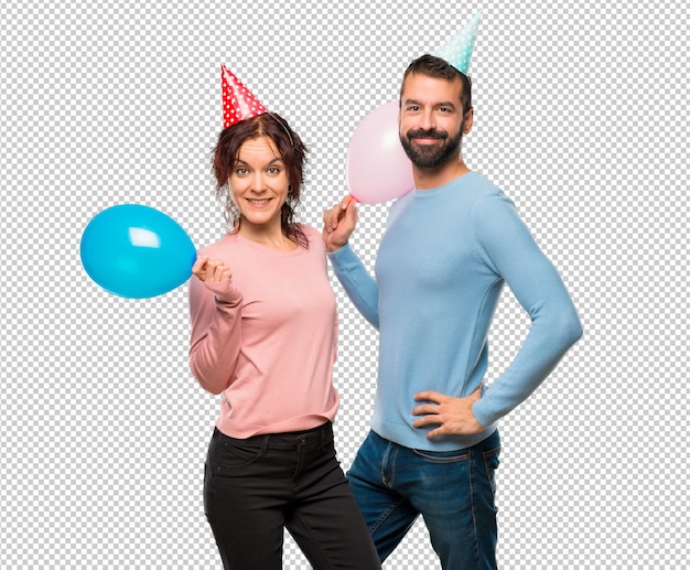 Pareja con globos y sombreros de cumpleaños posando con los brazos en la cadera y sonriendo