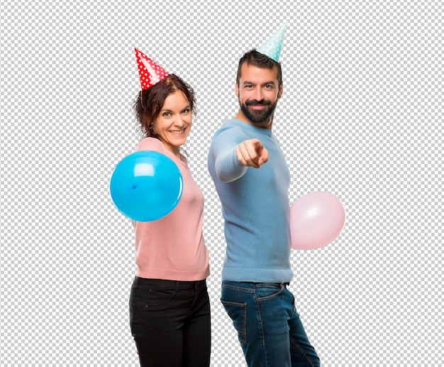 PSD pareja con globos y sombreros de cumpleaños puntos dedo con una expresión de confianza