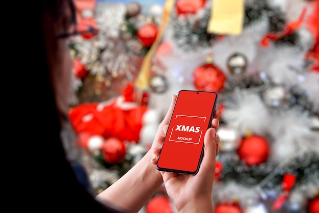 Ragazza con un telefono davanti a un albero di Natale. Display del telefono isolato per la promozione di app o messaggi di saluto